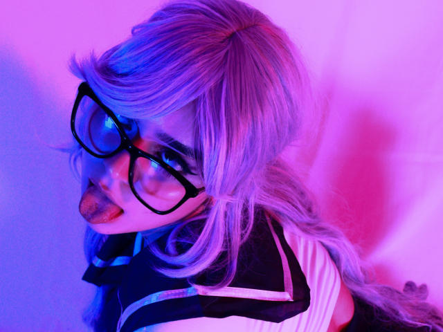 EvaSpicy, con cabello morado-azul, gafas negras y la lengua afuera, vestida con una blusa de marinero blanco y negro, bajo una luz rosa-morada neón, mostrando su lengua y una expresión juguetona..