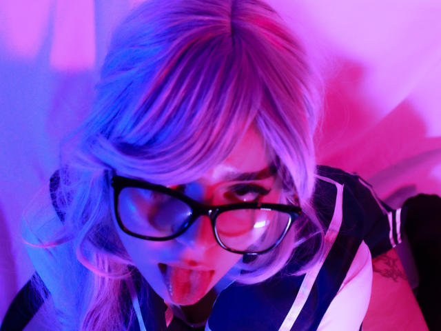 EvaSpicy, com cabelo roxo-azul, óculos pretos e língua de fora, vestindo um uniforme escolar, em uma foto com iluminação rosa e azul. Seu rosto está ligeiramente borrado..