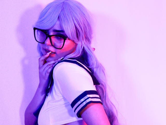 EvaSpicy, com cabelo roxo, óculos pretos e uma blusa branca de marinheira, mordendo o dedo, mostrando uma expressão fofa e tímida, com os seios parcialmente visíveis, sob iluminação roxa..