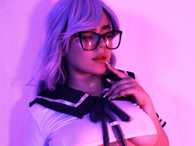 EvaSpicy, com o cabelo iluminado em tons de roxo, óculos pretos e uma blusa de marinheira branca com fita preta, mordendo o dedo, mostrando levemente os seios. Iluminação em tons de rosa e roxo..