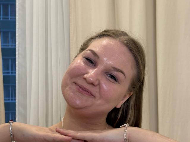 ValerieSchultheiss has shiny skin, a slight smile, and her hands are resting on her chest. She has brown hair, and a bracelet on each arm.
