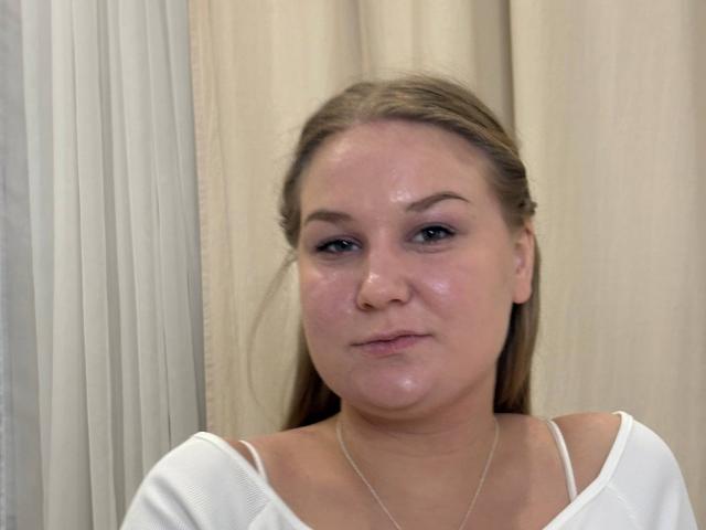 ValerieSchultheiss has shiny, flushed skin with a slight pout, blue eyes, and long brown hair. She wears a white top with thin straps, and a delicate necklace.