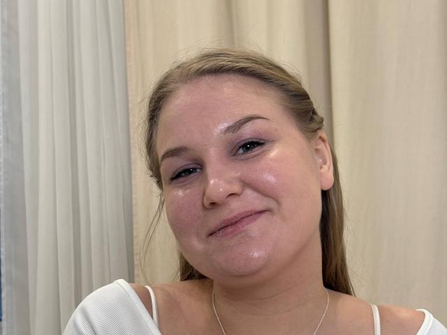 ValerieSchultheiss has shiny, slightly flushed skin, brown eyes, and a subtle smile. She's wearing a white top with thin straps, and her brown hair is loosely pulled back.