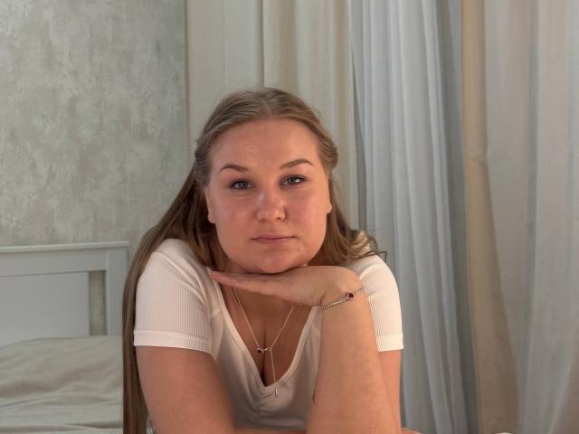 ValerieSchultheiss has long blonde hair, a white V-neck shirt, and rests her chin on her hand, looking at the camera with a neutral expression, showing her small tits.