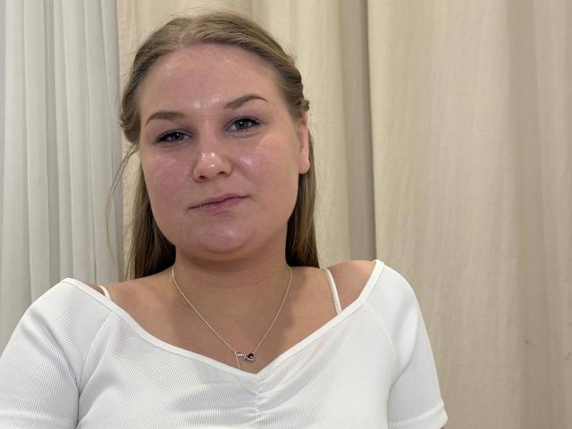 ValerieSchultheiss with a shiny face, slight pout, wearing a white top with thin straps, and a small necklace, has blonde hair and a neutral expression.
