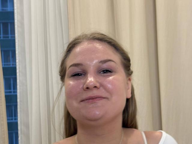 ValerieSchultheiss has shiny, glistening skin on her face, with light brown hair pulled back, and a slight, goofy smile, wearing a white top, looking directly at the camera.