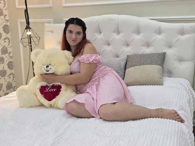 AnnaKimburli with red hair, pink off-shoulder dress, sitting on white bed, hugging beige teddy bear with red heart and "Love" text, on her left, legs spread.