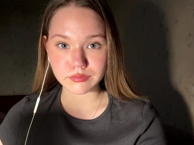 Juliet Erbst tem pele clara, olhos azuis, cabelo castanho liso, usa um piercing no nariz e está vestindo uma camisa cinza com um fone de ouvido branco no ouvido direito..