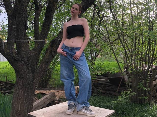 JulietErbst stands on a wooden plank, wearing a black strapless top, loose blue jeans, and gray sneakers, with her hands near her jeans, outside a tree-filled yard.