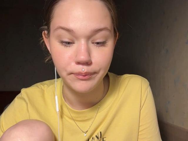 Juliet Erbst heeft een geel shirt, een neusring, en witte oordopjes, met een lichte glimlach, bruine ogen, en een kleine gouden ketting, en zit met de knieën opgetrokken..