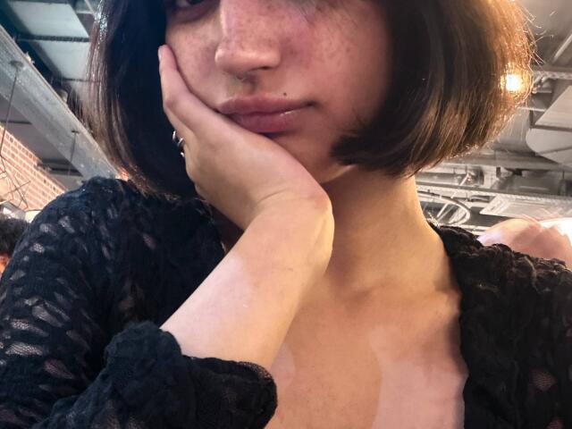 HeikeCarchi with a nose ring, freckled face, and short brown bob, touching her cheek with her hand, wearing a black lace top, showing a bit of her tits.