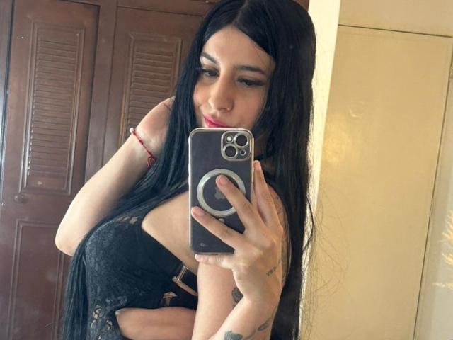 SamiSins, con lunghi capelli neri, indossa un reggiseno di pizzo nero, sta scattando un selfie allo specchio con la mano destra, mostrando i suoi seni e un braccialetto rosso, tenendo in mano un telefono con un anello..