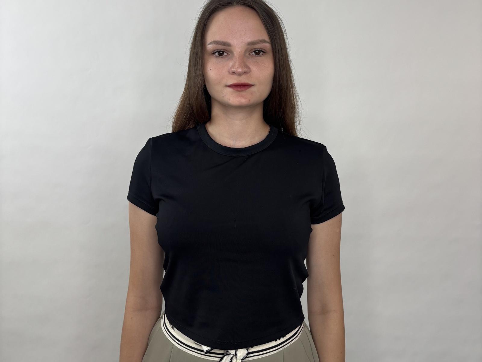 LoidaGess has a serious face, long brown hair, small tits, wearing a black short-sleeve shirt and beige pants, standing against a plain white wall.