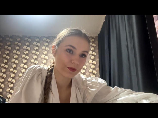 CaritaShover with braided blonde hair, wearing a white shirt, has red lipstick and a slight smile, sitting in front of dark curtains. Her tits are partially visible.