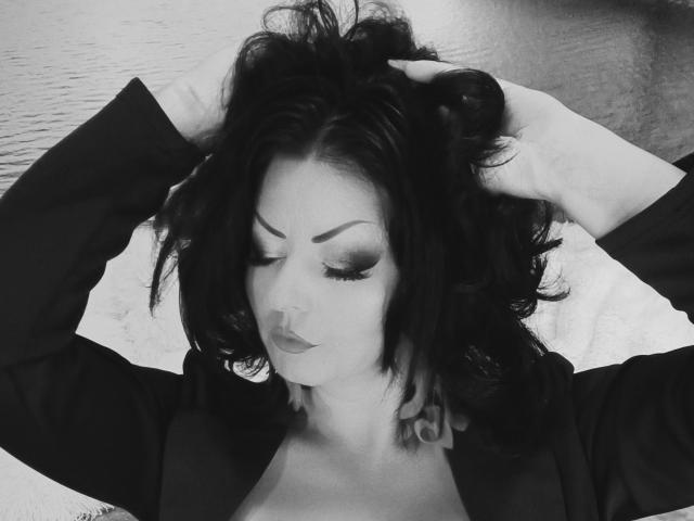 AmberSunshine mit dunklem, gewelltem Haar, den Händen im Haar, mit geschlossenen Augen, trägt ein schwarzes Oberteil, schwarzen Lippenstift und rauchigen Lidschatten und steht an einem Gewässer..