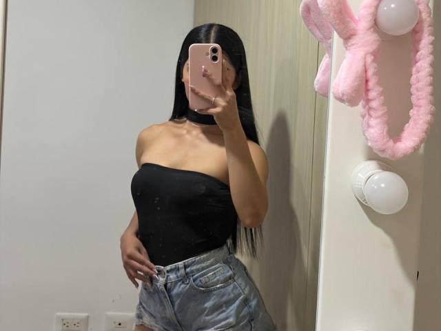 LaurentRay with long black hair, wearing a black strapless top and high-waisted denim shorts, taking a mirror selfie with a pink iPhone, showing her bare ass.