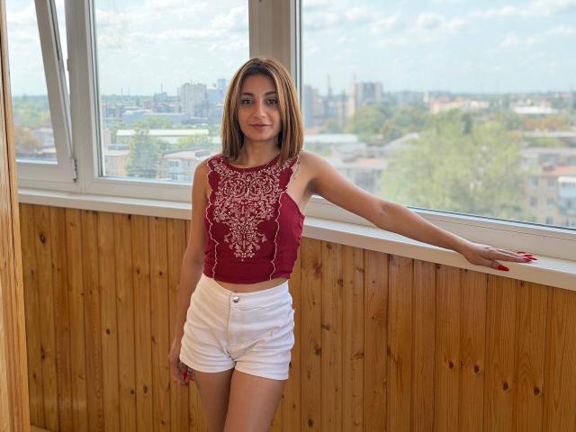 MiyaLinn with light brown hair, red embroidered sleeveless top, and white shorts, leaning on a window, wooden panel wall, city skyline visible, smiling, red nails, slender build.