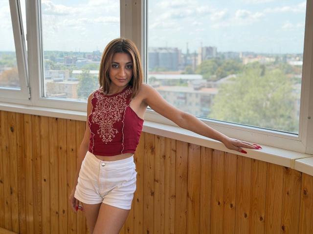 MiyaLinn with brown hair, red embroidered top, white shorts, leaning on window, wooden wall, red nails, smiling, standing, daylight, city view outside, right arm on window sill.