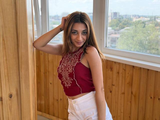 MiyaLinn with brown hair, red embroidered top, white pants, leaning against wooden wall, hand in hair, smiling, visible tattoo on arm, sunny window, city and trees outside.