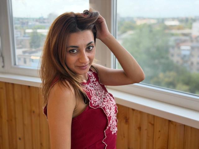MiyaLinn with brown hair, wearing a red top with white lace, smiling, hand in hair, looking at camera, standing by window, wooden wall, daytime.