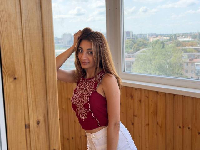 MiyaLinn with brown hair, red lace-up top, and white pants, standing by a wooden wall, hand in hair, looking at camera, city view through window in background.