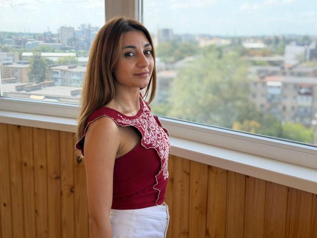 MiyaLinn with medium brown skin, long brown hair, red lace-up top, small tits, white pants, standing by window, wooden wall, smiling slightly, city view outside.
