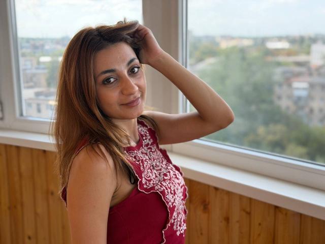 MiyaLinn has light brown skin, brown eyes, and long brown hair. She's wearing a red sleeveless top with white embroidery, smiling, and touching her head. Her tits are visible.