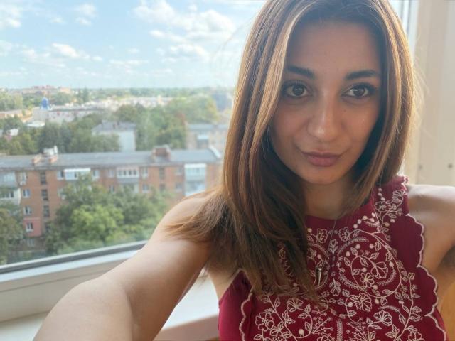 MiyaLinn with brown eyes, wearing a red sleeveless top with white embroidery, smiling slightly, brown hair, fair skin, taking a selfie near a window, looking directly at camera.