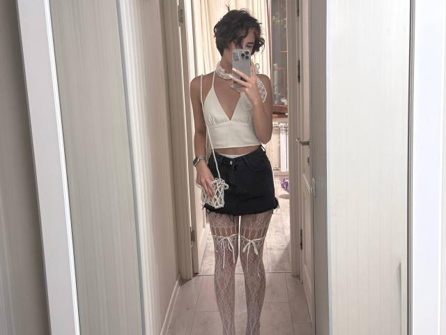 TameikaMacclellan with short curly hair, white crop top, black mini skirt, white lace thigh-highs, holding phone in mirror, wearing choker and wrist bracelet, standing in hallway.