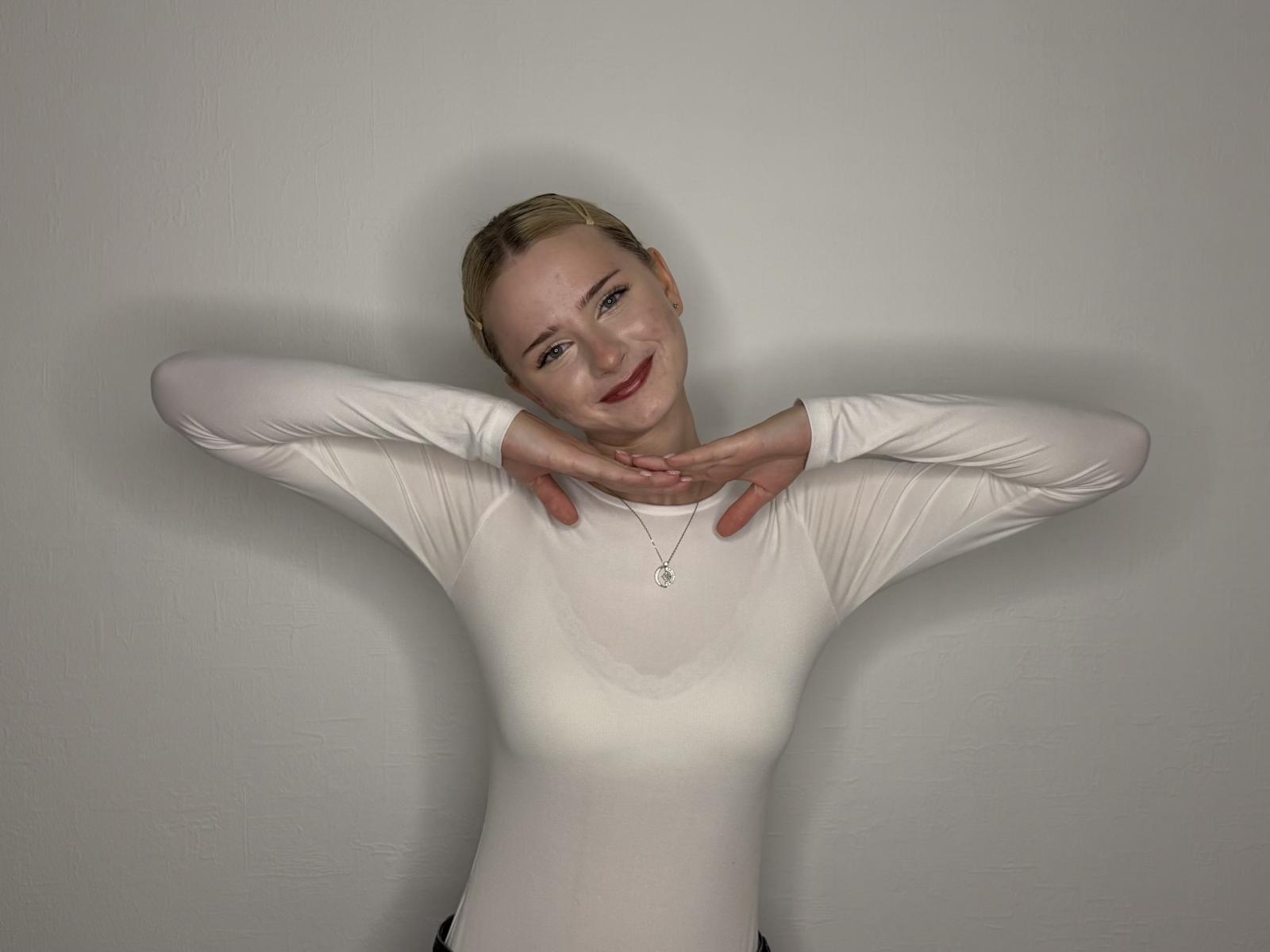 NeidaOhm has blonde hair, wearing a white long-sleeve shirt, arms raised, hands touching neck, smiling, small breasts, necklace, light skin, standing against a plain wall.