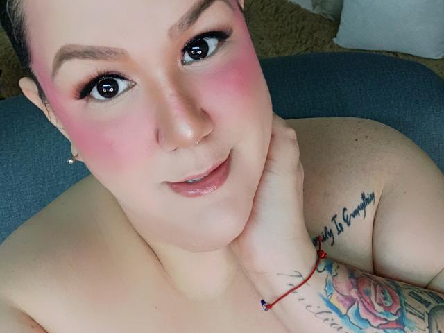 GiiannaMorrys with pink blush, looking at the camera, smiling, pink eyes, hand on chin, tattoos on arm, red bracelet, bare shoulders, short brown hair, and a nose ring.