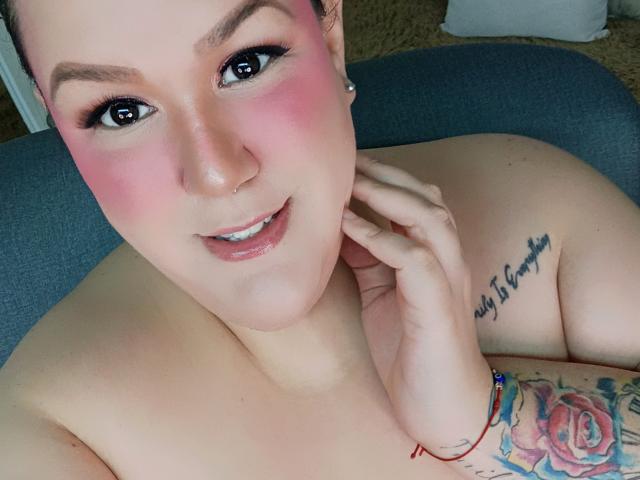 GiiannaMorrys with pink blush, black eyeliner, smiling, hand near neck, red bracelet, tattoo on right shoulder, and colorful rose tattoo on right arm. Topless, showing tits.