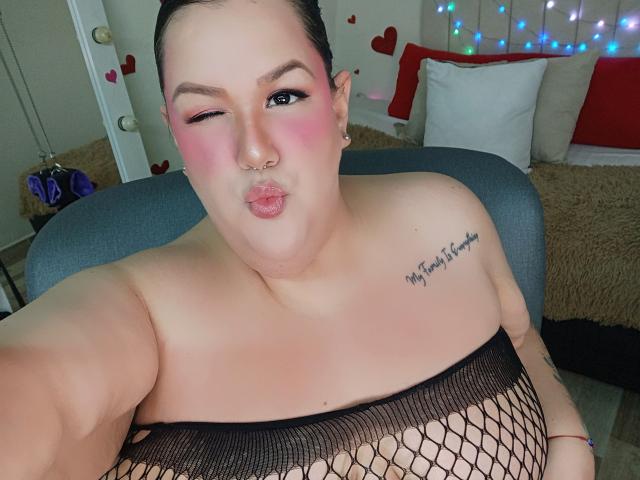 GiiannaMorrys with puffy pink cheeks, winking, wearing black fishnet top, showing big tits, tattoo on right shoulder, nose ring, and puckered lips, sitting in chair.