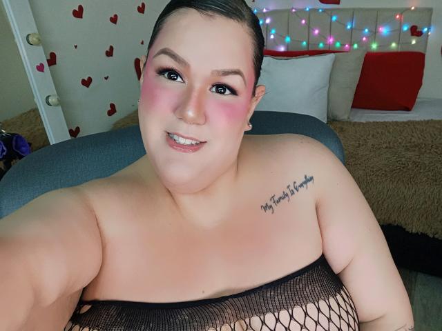 GiiannaMorrys with pink blush, black mesh top, visible tits, tattoo on right shoulder, smiling, short brown hair, small nose ring, colorful lights in background, sitting on gray bed.
