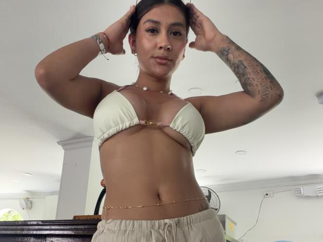 ThaliaAres with tan skin, small tits in a white bikini top, arms up, hands in hair, tattoo on left arm, gold chain, and a belly chain, standing in a room.