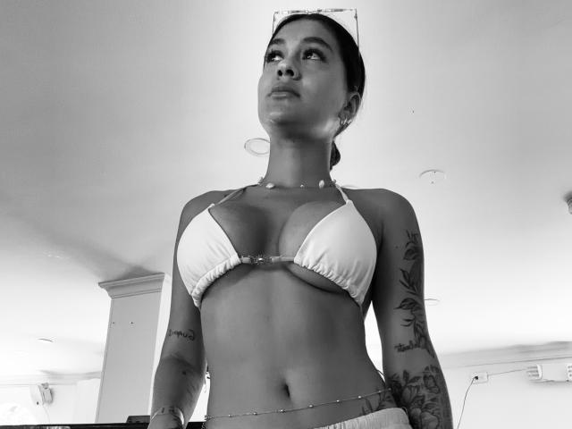 ThaliaAres with large boobs in a white bikini top, tattoos on arms, chain around waist, looking up, black and white photo, wearing a cap, low angle shot.