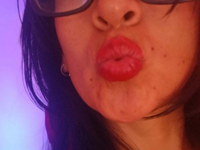 ZarayFeet's face with red lips puckered in a kiss, wearing glasses, black hair, and a silver ear ring, showing freckles and small moles. Purple-blue background.