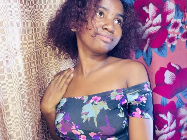 Yollande with curly brown hair, dark skin, wearing an off-shoulder floral top, has a hand on her chest, smiling, showing small tits, and a cute face.