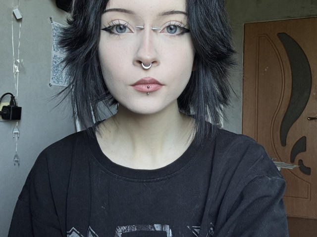 Liane Badenoch, com cabelo preto, piercing de prata no nariz, delineador preto, máscara de olhos prateada e piercing rosa nos lábios, vestindo uma camisa preta, olhando diretamente para a câmera..