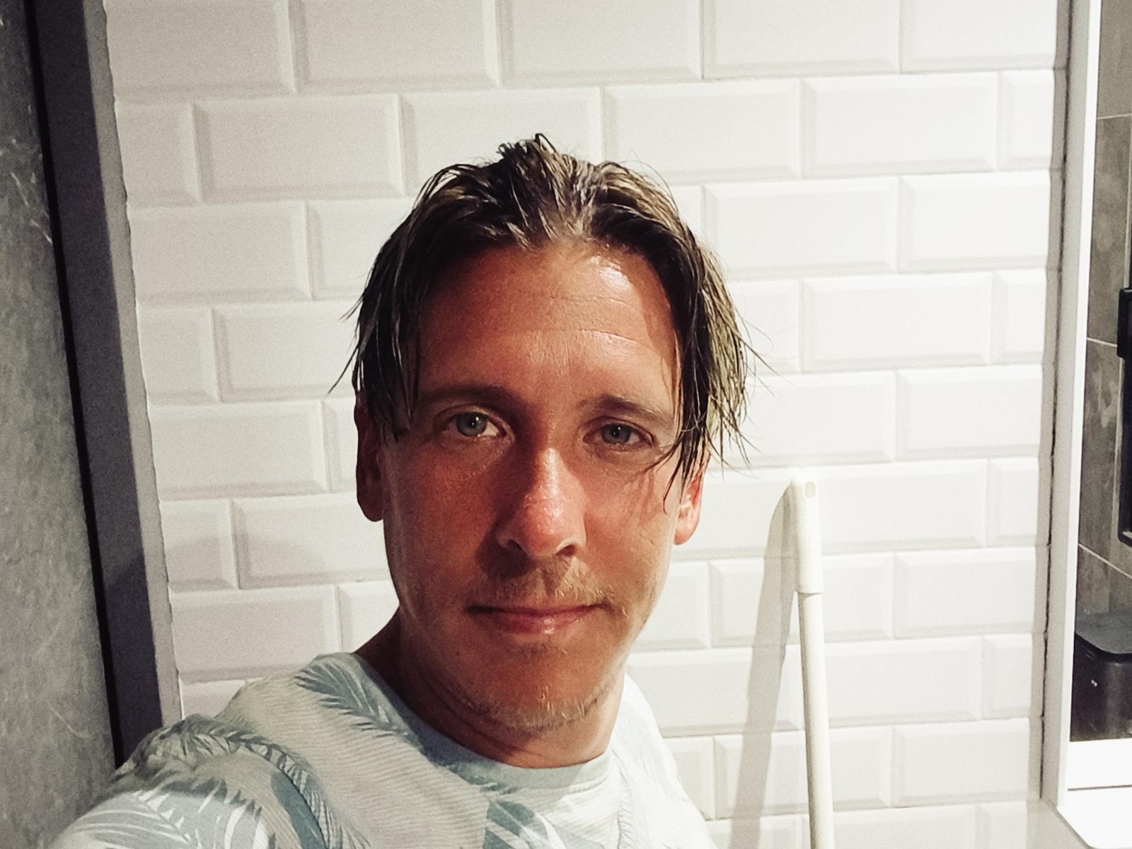 JefreyJoy, avec ses cheveux bruns mouillés, une légère barbe de quelques jours et un sourire discret, portant une chemise à motifs de feuilles vertes, dans une salle de bain aux carreaux blancs..