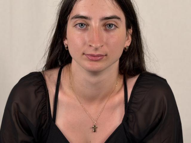 NicolHarp hat langes, dunkles Haar, blaue Augen und ein leicht rosafarbenes Gesicht. Sie trägt ein schwarzes, durchsichtiges Oberteil, kleine Ohrringe und eine goldene Kreuzkette. Ihre Brüste sind mittelgroß..