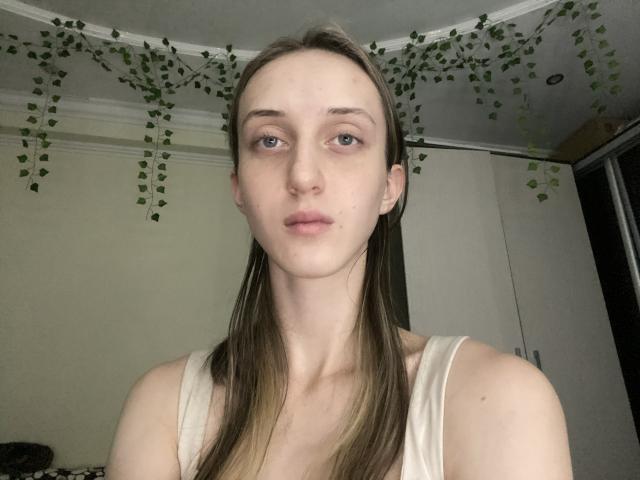 TwanaVanrossum tem pele clara, olhos azuis e cabelos castanhos longos, vestindo uma camiseta branca, com uma expressão neutra em um quarto com pouca luz, decorado com guirlandas de folhas verdes..