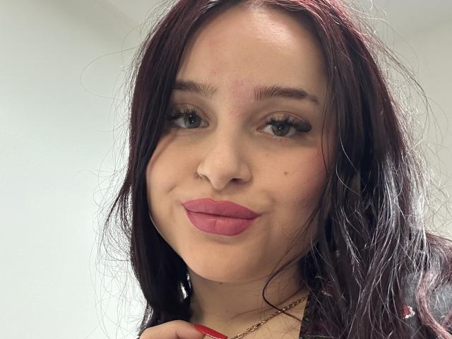 Luucie has dark red hair, green eyes, pink lipstick, and a small mole on her left cheek. Her red nails and gold necklace are visible. She's smiling slightly.