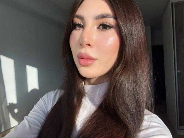 LeanoraMyree has long brown hair, full pink lips, and a shiny face with makeup. She's wearing a white top, and sunlight casts shadows on her.