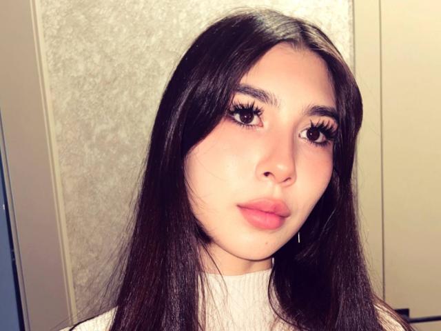 LeanoraMyree has long, straight black hair, full pink lips, and thick, long, dark eyelashes. She's wearing a white turtleneck and has a subtle, confident, sexy look.