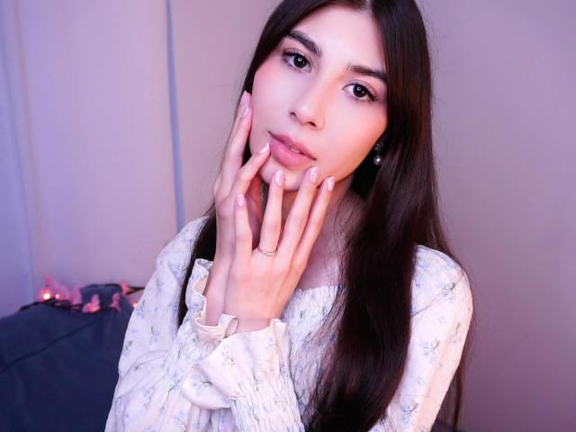 LeanoraMyree with long brown hair, fair skin, and pink lips, touching her face with her fingers, wearing a white floral dress, looking at the camera with a gentle expression.