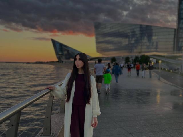 Leanora Myree, con el pelo largo y castaño, vestida con una bata blanca sobre un vestido oscuro, está de pie junto a una barandilla al atardecer. Se ven personas caminando al fondo, y agua a la izquierda..