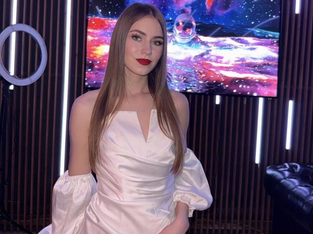 CaritaShover with long brown hair, red lipstick, wearing a white off-shoulder satin dress, standing in front of a colorful space-themed TV, ring light on the left.