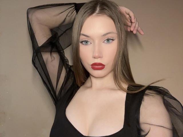 CaritaShover with long brown hair, blue eyes, and red lipstick, wearing a black sheer top, has her arm raised, exposing her boobs, and looking directly at the camera.