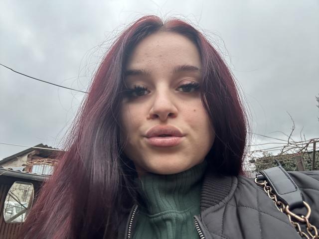 Luucie a les cheveux roux, des lèvres pulpeuses, et porte un pull vert à col roulé avec une veste matelassée grise. Elle a une chaîne autour du cou et l'arrière-plan montre un ciel légèrement nuageux..